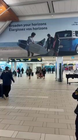 The media file depicts an interior scene within an airport terminal. The primary subjects are people, including travelers with luggage and a woman playing a grand piano. Objects visible include surfboards, luggage, a blue Volkswagen ID. Buzz van in a billboard advertisement, and airport signage. The setting is a brightly lit airport concourse with a tiled floor and high ceilings, suggesting it's daytime.

The activity taking place is the normal hustle and bustle of an airport: people are walking, presumably for flights, while a pianist provides live music, creating a pleasant ambiance. The overall emotion conveyed is one of travel and perhaps anticipation, with the music adding a touch of relaxation.

Notable details include the large billboard advertisement at the top, which reads "Broaden your horizons Élargissez vos horizons" and features the Volkswagen ID. Buzz. The airport signage indicates "Portes" (Gates) and "Tous les passagers" (All passengers), with bilingual text consistent with a Canadian location. The woman playing the piano appears focused and possibly enjoying her performance. A person in the foreground on the right is partially visible, looking towards the left. Another person in the foreground on the left is pulling a suitcase. The presence of a piano suggests an effort to enhance the passenger experience. The weather outside is not discernible from the interior view. The media file is filmed in Richmond, Canada.