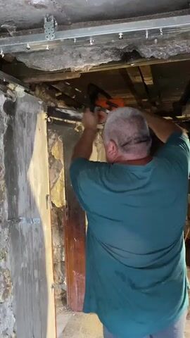 A person wearing a teal t-shirt and gray shorts stands with their back to the camera in a dimly lit, unfinished room. The walls are rough and appear to be made of concrete or stone, with visible damage and peeling plaster. Above, exposed wooden beams and structural elements are visible, along with metal framing and what appears to be insulation. The person is holding a chainsaw and appears to be preparing to cut something. The overall environment suggests a construction or demolition site.