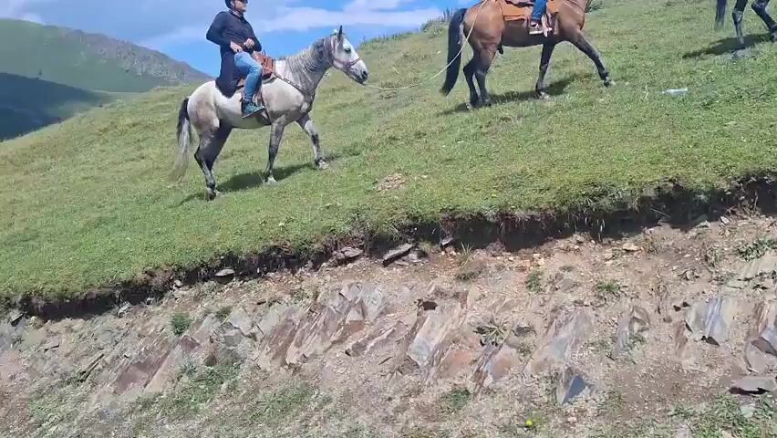 The video shows a group of people on horseback riding up a grassy incline under a bright blue sky with scattered white clouds. There are at least six horses and several riders, with some people also sitting or squatting on the grass at the side. The riders are wearing casual clothing and some have helmets. The horses are of various colors, including white, brown, and black. The environment appears to be a mountainous or hilly landscape with sparse vegetation and some rocks. The camera appears to be moving slightly, possibly from a moving vehicle or while the person filming is walking.