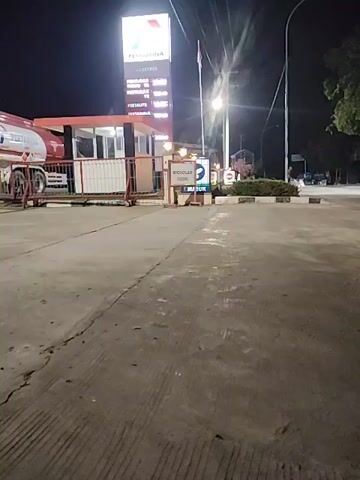 The video shows a nighttime view of a gas station. A tall sign with prices is illuminated. A truck is parked at the station. Traffic passes by on a road in front of the station. Several motorcycles are seen driving past the gas station entrance. One motorcycle with two riders passes directly in front of the camera. The ground appears to be concrete. The lighting is artificial, coming from streetlights and the gas station's illumination.