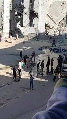 The video depicts a group of people, including several children and adults, in an outdoor area characterized by damaged buildings. The ground is uneven and appears to be a street or courtyard, with debris scattered around. Many individuals are holding or interacting with white plastic containers, some of which appear to be buckets. Several people are gathered around a central point where water is being dispensed, suggesting they are collecting water. Some are filling their containers, while others are carrying them away. The environment appears to be one of distress or scarcity, with the damaged infrastructure and the focus on water collection. The lighting suggests it is daytime. The people are dressed in casual clothing, with some wearing head coverings. The overall scene conveys a sense of community activity in a challenging environment.