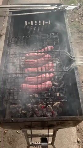 The video shows a close-up, overhead view of a barbecue grill with several sausages cooking on a rack over hot coals. Smoke is rising from the grill, obscuring some of the sausages. The grill appears to be a rectangular metal box with a stand. In the background, there are metal skewers. The ground is visible below the grill.