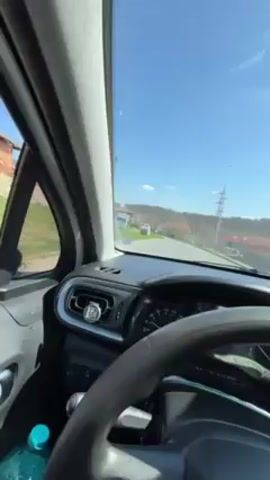 The video shows a first-person perspective from inside a car, looking out the windshield while the car is parked. The sky is blue, and the sun is shining. The car is parked on a street, and there are houses and trees visible in the distance. There is a water bottle sitting on the dashboard and an air freshener in the air vent. There is music playing.