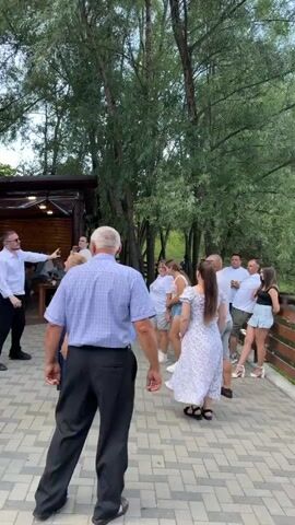 The video shows a group of people dancing outdoors at an event. In the foreground, an older man in a blue checkered shirt and black pants is dancing, facing away from the camera. To his left, a woman in a dark blue dress is also dancing. Behind them, a man in a white shirt and black pants, wearing sunglasses, is dancing with his arm raised. Further back, several other people are dancing, including women in dresses and men in casual wear. The setting appears to be a paved outdoor area surrounded by trees, with a wooden structure that looks like a covered seating area or restaurant in the background. The overall atmosphere suggests a celebration or party.