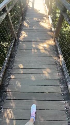 A first-person perspective video shows a person walking down a wooden elevated walkway. The walkway is surrounded by railings made of wood and metal mesh. Sunlight filters through the trees, casting shadows on the wooden planks. The person is wearing light-colored pants and white sneakers with pink and blue accents. The walkway appears to be in a natural environment with lush green foliage visible on either side. The person is the only visible subject.