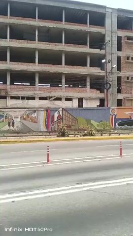 The video, shot from a moving vehicle, shows an unfinished, multi-story building under construction. The building is predominantly brick and concrete, with many open spaces where windows and walls would be. In front of the building, a roadside barrier painted with murals is visible. Cars drive by on the road. A few people can be seen walking on the sidewalk beside the road. The weather appears to be sunny. The video is slightly shaky, likely due to the movement of the car.