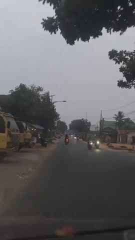 The video shows a view from inside a vehicle looking forward along a paved road. The sky is overcast and appears to be dusk or dawn. On the left side of the road, there are trees, roadside stalls with awnings, and several parked motorcycles. A yellow truck is also visible. On the right side of the road, there are more trees, some buildings, and parked trucks and vehicles. Several motorcycles are driving along the road in the same direction as the viewer. The overall impression is a rural or semi-urban road scene in low light conditions.