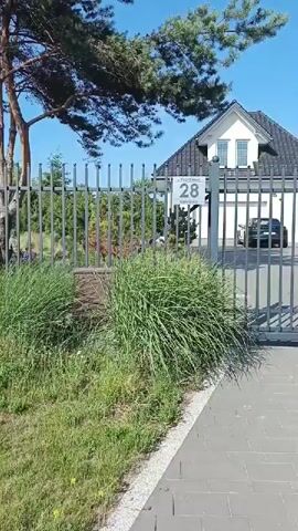 The video pans across a modern, two-story white house with a dark shingled roof. A tall gray metal fence runs along the front of the property, with a gate in the foreground. A sign on the gate reads "28". A car is visible in the driveway behind the fence. Lush green grass and ornamental plants are growing in front of the fence. The sky is clear and blue. The video appears to be taken on a sunny day.