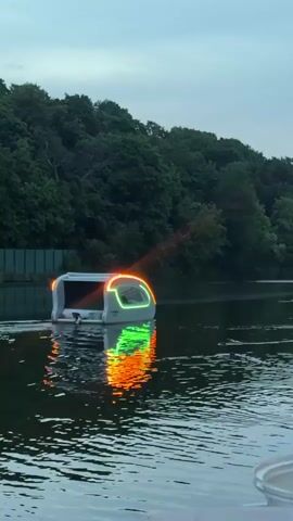 A remote-controlled vehicle with bright orange and green lights cruises on the water. Trees line the background, reflecting the lights on the water's surface. The sky is overcast and dim, indicating it is either dusk or dawn.