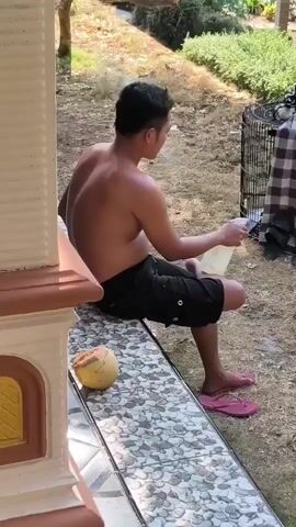 A shirtless man in black shorts and pink flip-flops sits on a tiled porch next to two bird cages in a grassy yard. One cage is covered with a brown and white checkered cloth. The man is holding a clear plastic bottle with a white lid and appears to be spraying something from it. A coconut rests on the porch. The man then stands up and approaches the covered cage, still holding the bottle. The video pans to show both cages and the surrounding yard with dry grass and some greenery in the background.