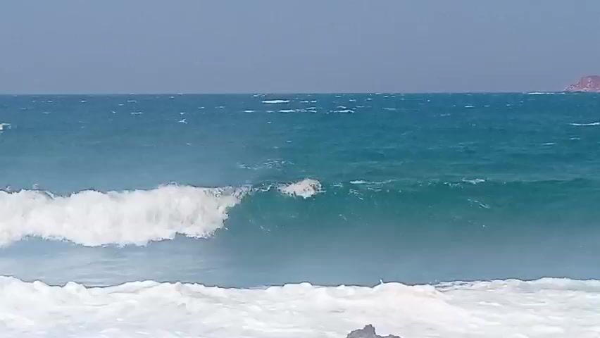 The video captures a wide shot of the ocean with choppy, turquoise waves crashing onto a rocky shore. A line of surfers, each on a surfboard, are visible in the middle ground, paddling in the water. They are positioned to the left of the frame and are spaced out, with some appearing closer together than others. The sky is clear and hazy, with a distant landmass or island visible on the horizon. The water is foamy and turbulent, indicating strong currents or wind.