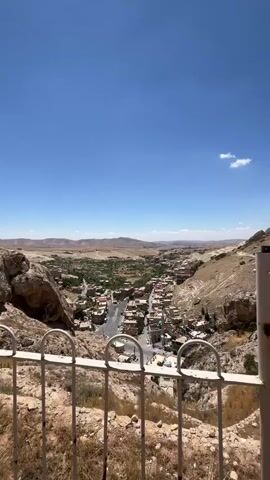 The video shows an elevated view of a town, framed by a low white metal fence with rounded tops. The town, nestled in a valley, consists of closely packed buildings with red tile roofs, intersected by roads. Beyond the town, the landscape extends into a flat, arid plain under a bright blue sky with a few scattered clouds. The foreground features rocky, dry terrain with sparse vegetation. To the right, a large rock formation rises, adding to the rugged character of the scene.