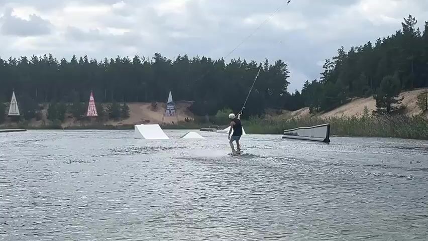 A person is wakeboarding on a lake. They are wearing a helmet and a dark-colored shirt and shorts. The person is being pulled by a cable system. There are several ramps and obstacles in the water, including a large white ramp on the left and a smaller white ramp in the center. In the background, there is a line of trees, a sandy hill, and a cloudy sky. There are also some decorative triangular structures on the hill. The water is choppy and there is a wake behind the wakeboarder.