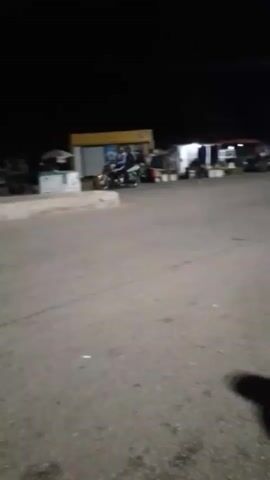 A dark, grainy video shows a street scene at night. The camera is moving, resulting in a blurry image. In the foreground, there is a paved surface, possibly a road or parking lot. In the background, there are several dimly lit buildings or stalls, and a few motorcycles are visible, some with riders. The lighting suggests artificial illumination from streetlights or the establishments themselves. The overall impression is that of a somewhat busy, albeit dark, outdoor commercial area at night.