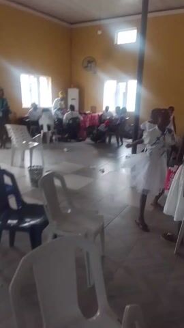 The video shows a gathering of people in a room. Most of the adults are seated at tables in the center, while a few children are walking around and appear to be dancing.  Most people in the video are wearing white clothing. The room is decorated in a simple fashion, with a tiled floor, yellow walls, and several windows. A couple of ceiling fans are also visible. Several chairs, both empty and occupied, are present throughout the room. There is a column present in the lower-right of the frame.