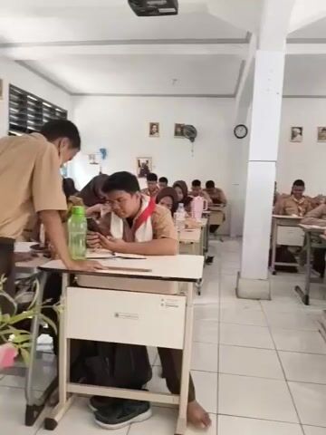 The video shows a classroom setting with rows of desks. Several students are seated at the desks, some of whom are wearing uniforms and headscarves. Some students appear to be looking at their phones, while others are engaged with papers or books. A person in a brown uniform with a red sash is visible in the foreground, looking down at their phone. Another student in the foreground, also in uniform, is leaning over their desk and possibly interacting with the student next to them. The classroom is well-lit with fluorescent lights. There are pictures and a clock on the walls.