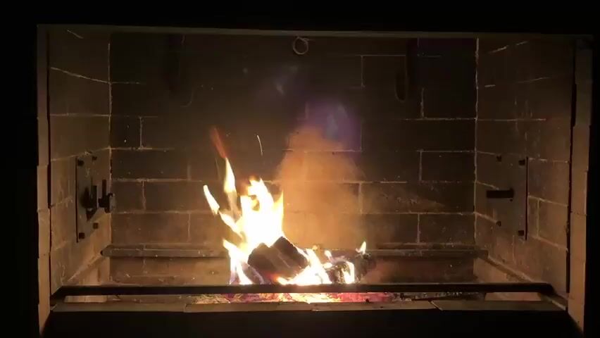 A fireplace is shown with a fire burning. The fireplace is made of brick. There are logs burning in the fire. There is some smoke rising from the fire. The flames are bright yellow and orange. There is a hint of blue flame at the top of the fire. There is a metal grate in front of the fireplace.