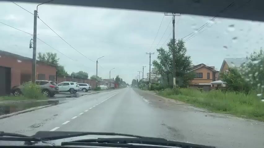 The video shows a view from inside a car driving down a wet, asphalt road. It appears to be a cloudy day, and rain is visible on the windshield. On the left side of the road, there are several garage buildings and parked cars. On the right side, there are houses and trees. The road is relatively straight and appears to be in a semi-rural or suburban area. The car is moving forward, and the scenery is passing by. There are power lines and poles running along the side of the road.