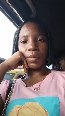 A young woman with dark skin and braided hair is in the foreground of the video, looking directly at the camera. She is wearing a light pink t-shirt and a silver necklace with a distinctive "V" shaped pendant. Her right hand is propped up on her chin, and her gaze is steady. In the background, slightly out of focus, another person, also appearing to be a woman with dark skin and braided hair, is visible in the back seat of what appears to be a vehicle. The environment outside the vehicle suggests it is daytime, with hints of buildings and structures visible through the window. The camera appears to be held by the young woman in the foreground, indicating a selfie-style recording. The overall mood is neutral, with the woman in the foreground appearing pensive or contemplative.