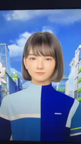 A video shows a digital rendering of a young woman with short brown hair and bangs, wearing a color-blocked blue and white top with "AISIN" on the chest. She is positioned against a backdrop of a modern city with clear blue skies. A small, cartoonish, white, winged creature floats in the scene. The video is slightly shaky, as if recorded on a phone pointing at a screen. The woman looks down and then the video focuses on the cartoon creature.