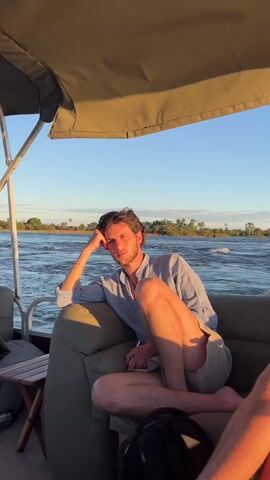 A young man is sitting on a boat on a body of water, appearing to be a river, and looking toward the camera. He is fair-skinned with dark curly hair and a beard. He is wearing a light blue long-sleeved button-down shirt and khaki shorts. He is wearing a necklace and a bracelet on his left wrist. His head is resting on his left hand and his legs are crossed. The background shows the river with trees and vegetation on the banks, and a clear blue sky. The boat is moving, as evidenced by the water flowing past. The overall light suggests late afternoon or early evening.