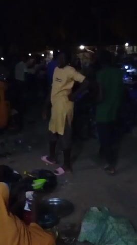The video depicts a nighttime scene outdoors with several people present. The lighting is dim, but there are some overhead lights illuminating the area. A person wearing a yellow tunic and pink sandals is prominently visible and moves through the crowd. Other individuals wear clothing in colors like green, blue, and dark shades.  The ground appears to be paved and cluttered with objects, including basins, containers, bags, and scattered debris. People seem to be interacting and milling about in a casual manner.