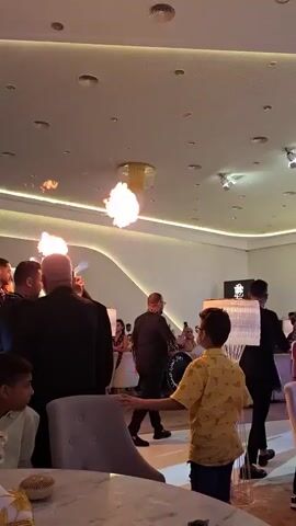 The video shows a group of people inside a brightly lit, modern event space. In the foreground, a young person in a yellow patterned shirt stands facing away from the camera, looking towards a performance area. In the mid-ground, several men in dark suits are engaged in a fire-breathing performance. Flames are seen shooting from their mouths upwards towards the ceiling. Other guests are visible in the background, seated at tables and watching the performance. The space features a high ceiling with recessed lighting and decorative elements.