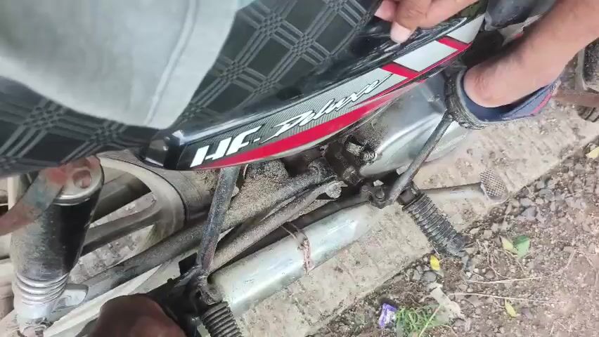 This is a close-up, low-angle shot of the side of a motorcycle. The seat, which is black with a diamond pattern, has "HF Deluxe" written on its side in white and red lettering. A person's hand is visible near the top of the seat, and their legs are positioned on the motorcycle. One leg, wearing a blue jean, is towards the back of the bike, while the other leg, wearing a dark blue sandal with red and white stripes, is resting on the kick-start lever. The motorcycle appears to be older and slightly dirty, with visible mud and dust on its components, including the engine, exhaust pipe, and rear wheel. The ground beneath the motorcycle is rough and appears to be made of dirt and small stones. The lighting suggests it is daytime outdoors.