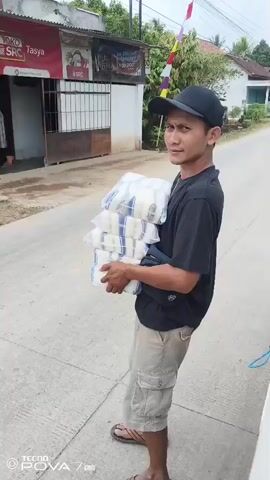 A man in a black t-shirt, tan cargo shorts, a black baseball cap, and flip-flops is standing on a paved road, carrying a stack of white items wrapped in plastic bags. He is holding the stack with both hands in front of him. The man is looking to his right, with a slight smile. Behind him, there is a building with signs in Indonesian. The road appears to be in a rural or suburban area, with trees and houses visible in the background. There are colorful flags hanging on poles in the distance. The video is recorded from a side-view perspective.