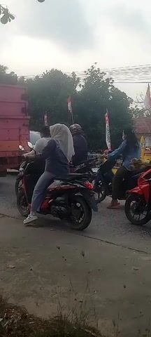 The video shows a street scene with several motorcycles and a truck. People are riding on the motorcycles, some with passengers. There are also flags visible, suggesting some kind of event or celebration. The environment appears to be a road with buildings and trees in the background. The lighting suggests it might be daytime, possibly overcast.