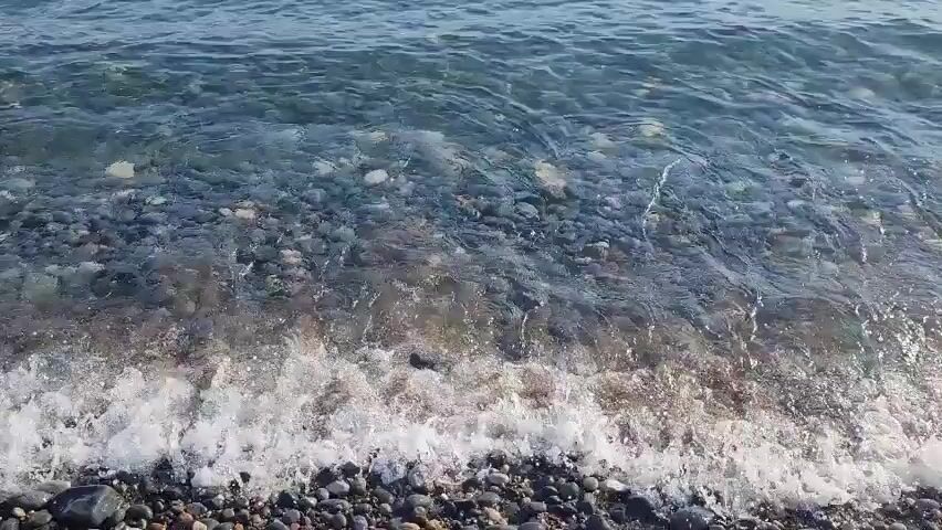 The video shows a close-up view of waves gently washing over a pebble beach. The water is clear, allowing the pebbles beneath to be visible. The pebbles are varied in size and color, ranging from dark gray to lighter brown and reddish hues. Small white foam crests form as the waves break on the shore. The camera angle is low, looking down at the water's edge. There are no people or animals visible in the frame.