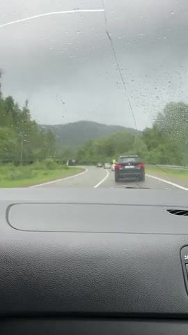 The video shows a view from inside a car looking out through a windshield with raindrops and a crack. The car is driving on a road with a black SUV in front of it. There are trees and hills in the background. The sky is cloudy and gray. The road appears to be a two-lane highway. The overall impression is that of driving in rainy weather through a rural or mountainous area.