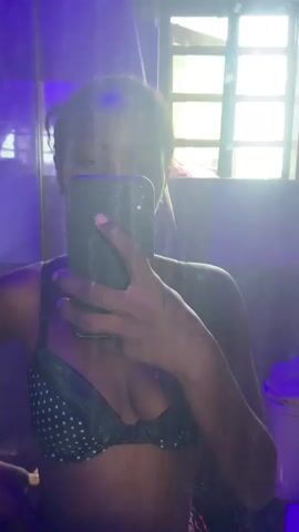 A person stands in a bathroom, holding a phone in front of their face to take a selfie in the mirror. The individual is wearing a black bra with white polka dots and a gold necklace with a wing pendant. The bathroom is illuminated with a purple light, and there is a window with a grid pattern behind the person. The walls of the bathroom appear to have a dark, shiny tiled surface. A canister sits in the periphery to the right. The focus is mainly on the person taking the photo, with the phone obscuring their face.
