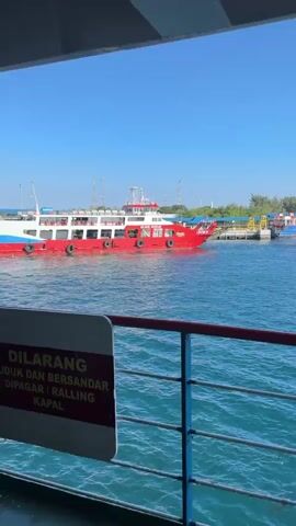 The video captures a view from a moving vessel, looking towards a red and white ferry docked at a pier. The ferry is a substantial size, with multiple decks and a white superstructure. It is painted red along its lower hull, with a blue stripe. The ferry appears to be at a port or harbor, with a clear blue sky overhead and calm, blue water. In the background, there are structures on the pier, including yellow cranes or lifting equipment, and some buildings. Further in the distance, mountains are visible under the clear sky. The camera perspective suggests the viewer is on another boat, likely also a ferry or similar transport, as indicated by the railing in the foreground. No people are clearly visible on the ferry or the pier. The scene suggests a typical day at a port.