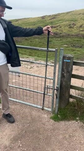 A person wearing a black jacket, white t-shirt, tan pants, and dark sneakers opens a metal gate by lifting a vertical bar attached to the gate and rotating it to the side. The gate is made of silver metal bars and has a mesh infill. To the right of the gate is a wooden post with a blue sign. The ground is a dirt path with small stones. In the background, there is a grassy hill with some purple heather. The sky is overcast. After opening the gate, the video shows a close-up of the person's legs as they walk forward on the dirt path.