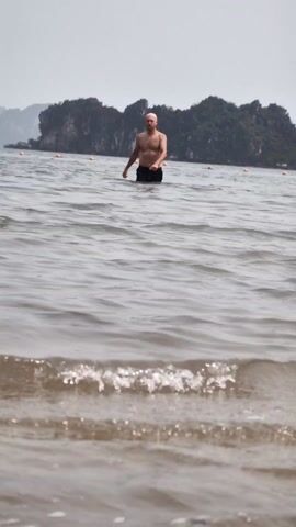 A medium shot shows a shirtless bald man in black swim trunks walking through the shallow, wavy water towards the viewer on a cloudy day.  The water is up to his mid-thigh.  There are small waves in the immediate foreground.  There are orange floats strung across the water in the middle ground. In the background, there is an island with rocky cliffs covered in dense green vegetation.