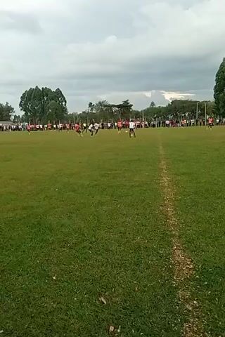 On a slightly overcast day, a soccer game is in progress on a lush green field. A small crowd of spectators lines the far edge of the field, their figures appearing as silhouettes against the backdrop of trees. Several players, wearing a mix of black, white, and orange jerseys, are actively engaged in the game, moving across the field as the ball is in play. A prominent white line is visible on the ground, running horizontally across the frame. The camera is handheld, and the person recording the scene is standing near the sidelines, with a portion of their arm and clothing visible in the foreground.
