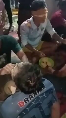 A group of people are gathered around a person lying on the ground, with some applying a white substance to the person's head and face. The person on the ground appears to be a child, and they are wearing a blue and black t-shirt with the word "DRAXON" visible. The people around the child are wearing casual clothing. The ground is covered with a patterned rug. The lighting is dim, suggesting it may be indoors or at night.