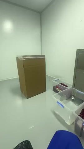 A room with white walls and a light gray floor. In the foreground, a person's legs and torso, wearing blue clothing, are visible. To the left, a large cardboard box stands upright. In the center and right of the frame, several clear plastic bins filled with red, bean-like objects are arranged on the floor. Behind the bins is a stainless steel machine with a hopper on top, from which the red objects are being dispensed into a clear plastic bin placed in front of it. A computer monitor is attached to the machine, displaying a green interface with graphs. A red indicator light is illuminated on the machine.