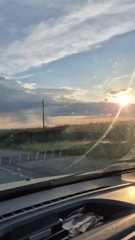 This is a video captured from inside a vehicle, looking out through the windshield. The scene is a rural landscape at sunset, with the sun low on the horizon, casting a warm glow. The sky is a mix of blue and dramatic clouds, with streaks of light breaking through. In the distance, there are fields of grass and some trees, with a single utility pole standing tall. A road, possibly a crossroads, is visible in the foreground, with a barrier of metal posts. The dashboard of the vehicle is visible at the bottom of the frame, with some crumpled paper in a storage compartment. The light filtering through the windshield creates a lens flare effect, with small circular reflections visible on the glass. The overall mood is serene and contemplative, evoking the beauty of a natural landscape at the end of the day. The location is identified as Eleshnitsa, Bulgaria, which helps to place this scene within a specific geographical context. The time of day is clearly sunset. The weather appears to be fair with some cloud cover, but the sun is still visible. There are no people visible in the video, and no discernible text.