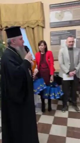 A group of people are standing in a room. On the left is a man dressed in a black robe and a black headdress, speaking into a microphone. Next to him is a woman in a red jacket who is holding several blue bags. To the right is a man in a grey blazer, and next to him is a man in a black suit with a black tie. All are standing on a checkerboard tiled floor. On the wall behind them are large framed posters with diagrams and descriptions in a foreign language. A Christmas tree is visible on the left side of the frame.
