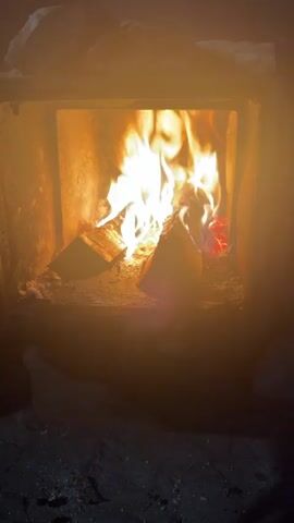A fireplace contains burning logs, with flames flickering brightly. The fireplace appears to be made of rough stone or brick. The surrounding area is dark and indistinct, suggesting a low-light environment.