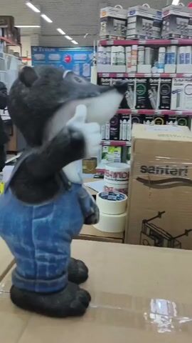 The video shows a statue of a raccoon wearing blue overalls, standing on a cardboard box in what appears to be a store. The raccoon statue is giving a thumbs-up gesture. In the background, several men are visible, and shelves stocked with various items can be seen, suggesting a retail environment, possibly a hardware or home improvement store. The lighting suggests an indoor setting with artificial lights.