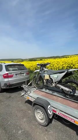 A static, eye-level shot features a grey dirt bike secured on a small trailer hitched to a silver BMW SUV. The dirt bike has the number 365 displayed on it. The trailer has a maximum speed indicator of 100. The background reveals a vibrant yellow field of rapeseed flowers extending to the horizon under a blue sky with scattered clouds. The vehicle is parked on a paved road. The overall environment suggests a rural or agricultural setting.