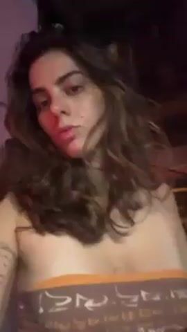 The video shows a woman with long, curly brown hair lying on the ground. She is wearing a patterned crop top. Several people are standing around her, their legs and torsos visible but faces out of frame. The lighting in the room alternates between red, blue, and purple, suggesting a party or club setting. The floor appears to be concrete or tile. The camera angle is mostly overhead.