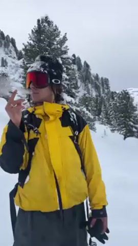The video shows a snowy mountain scene. In the first frame, a person wearing a yellow jacket, black hat and ski goggles is standing in the snow, smoking. In the background are snow-covered trees and a mountain. The next frames show a view of a ski slope with other skiers and snowboarders. There are trees, a ski lift, and buildings visible in the distance. The sky is overcast.