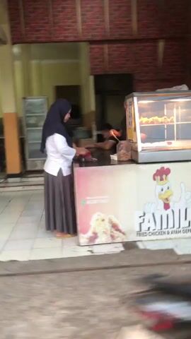A young woman wearing a hijab and a white top with a long grey skirt stands behind a counter in what appears to be a food establishment. She is facing away from the camera and interacting with someone on the other side of the counter, whose face is obscured. To her right, a display case filled with fried chicken is visible. The establishment has a brick-like wall in the background and tiled flooring. The overall scene suggests a transaction or service taking place at a food counter.