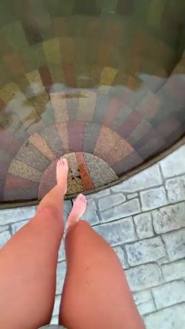 The video shows a person's legs as they stand on stone steps leading into a shallow pool of water. The steps are made of multi-colored stone tiles. The water is clear enough to see the steps underneath. The person is standing with their feet submerged, and the water is splashing slightly. The camera appears to be held by the person standing in the water, providing a first-person perspective. The surrounding area appears to be a tiled patio.