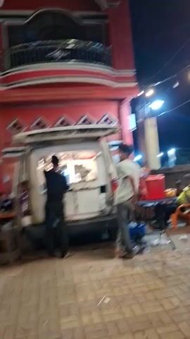 The video opens with a dimly lit scene at night, showing the back of a white van that appears to be a food truck, with its rear door open and illuminated from within. Two figures are visible near the van; one, wearing dark clothing, is standing in front of the open door, possibly working, while another person, wearing lighter clothing, is standing to the right of the van. A red building with decorative elements is visible in the background. The camera then pans rapidly to the left, showing what appears to be a street or outdoor area with various stalls and parked motorcycles illuminated by artificial lights, creating a blurred, motion-heavy effect. The scene then shifts to a dark road at night, with the headlights of oncoming vehicles creating bright streaks of light. Further down the road, more illuminated stalls or small shops are visible, and another motorcycle passes by, blurred by its speed. The overall impression is a street scene at night, likely a marketplace or food stall area, with ongoing activity and vehicle movement.
