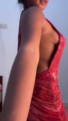 A woman is modeling a red floral halter dress with a low back, in what appears to be a bedroom. She turns to show the back of the dress, then places her hands on her hips. The room has plain white walls with a power outlet visible and a bed is partially seen in the background. A piece of electronic equipment with a charging cable is visible on the bed stand. The woman has her hair up in a bun.